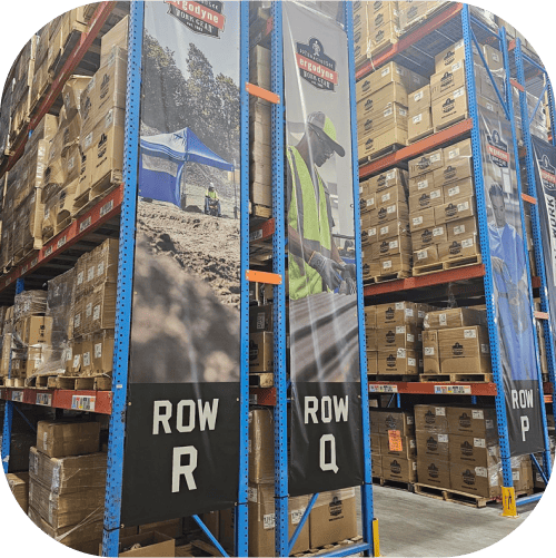 Photograph of Ergodyne's fullfillment center interior with high shelving units stocked with cardboard boxes. The shelves are labeled with 'ROW R,' 'ROW Q,' and 'ROW P' respectively, indicating different storage areas or aisles within the warehouse.																On the side of each shelving unit, there are vertical banners featuring the Ergodyne brand and photos. One banner appears to show a worker in a high-visibility vest, suggesting a focus on workplace safety and equipment. The environment is organized, and is a large-scale storage system typical of distribution centers or large retail backrooms.