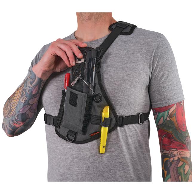 Tattooed person wearing gray shirt and black chest harness holding tools, including a red pocket knife, keys and a yellow tool; hand adjusting shoulder strap