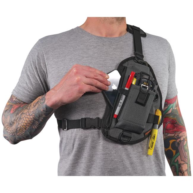 Tattooed person's torso in gray shirt wearing a black chest sling bag; hand placing a smartphone into a pocket holding pens and tools.