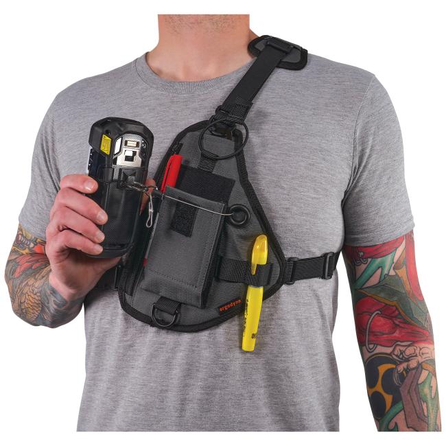 Torso of person in gray shirt wearing black shoulder harness with pouch, holding a handheld device; yellow pen and tattooed forearms.