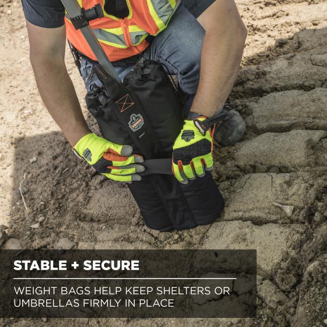 Person in a high-visibility vest and neon gloves kneeling on dirt, holding a black weight bag strapped around a pole.