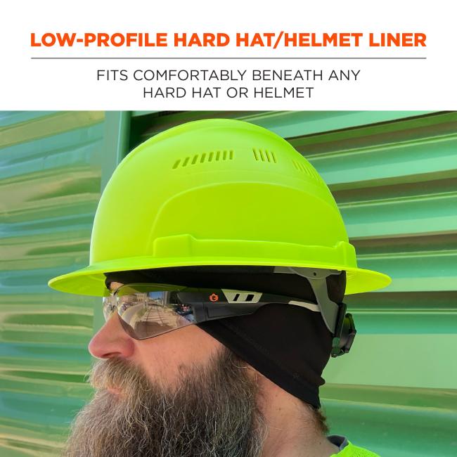 Side profile of a bearded man wearing a neon yellow hard hat, safety glasses and a black helmet liner against green corrugated metal background.