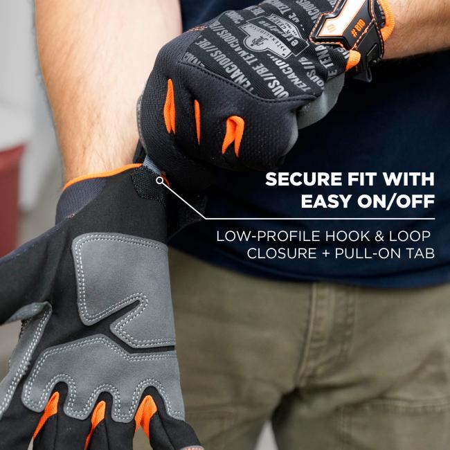 Close-up of black and gray work gloves with orange accents being pulled onto a person's hand, showing hook-and-loop closure and printed text