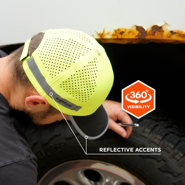 Close-up of person wearing neon perforated safety cap with reflective stripe, leaning over a tire beneath a rusted metal edge
