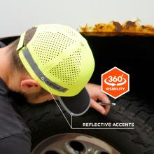 Close-up of person wearing neon perforated safety cap with reflective stripe, leaning over a tire beneath a rusted metal edge